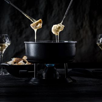 Zwei Stücke Brot eingetaucht in heisses Fäden ziehendes Käsefondue vom Chäsbueb und im Hintergrund Gläser Weisswein. Passend zum Fondue.